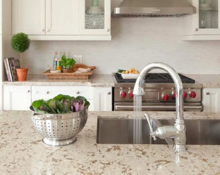 Kitchen sink and running faucet set in a spotted beige countertop with a colander of leafy greens.